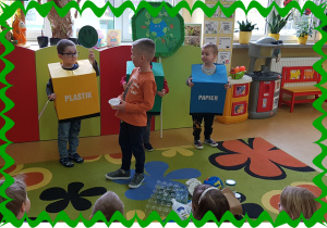 Trzech chłopców w kolorowych , kartonowych pudłach z napisami :plastik, szkło, papier. Chłopiec segreguje śmieci.